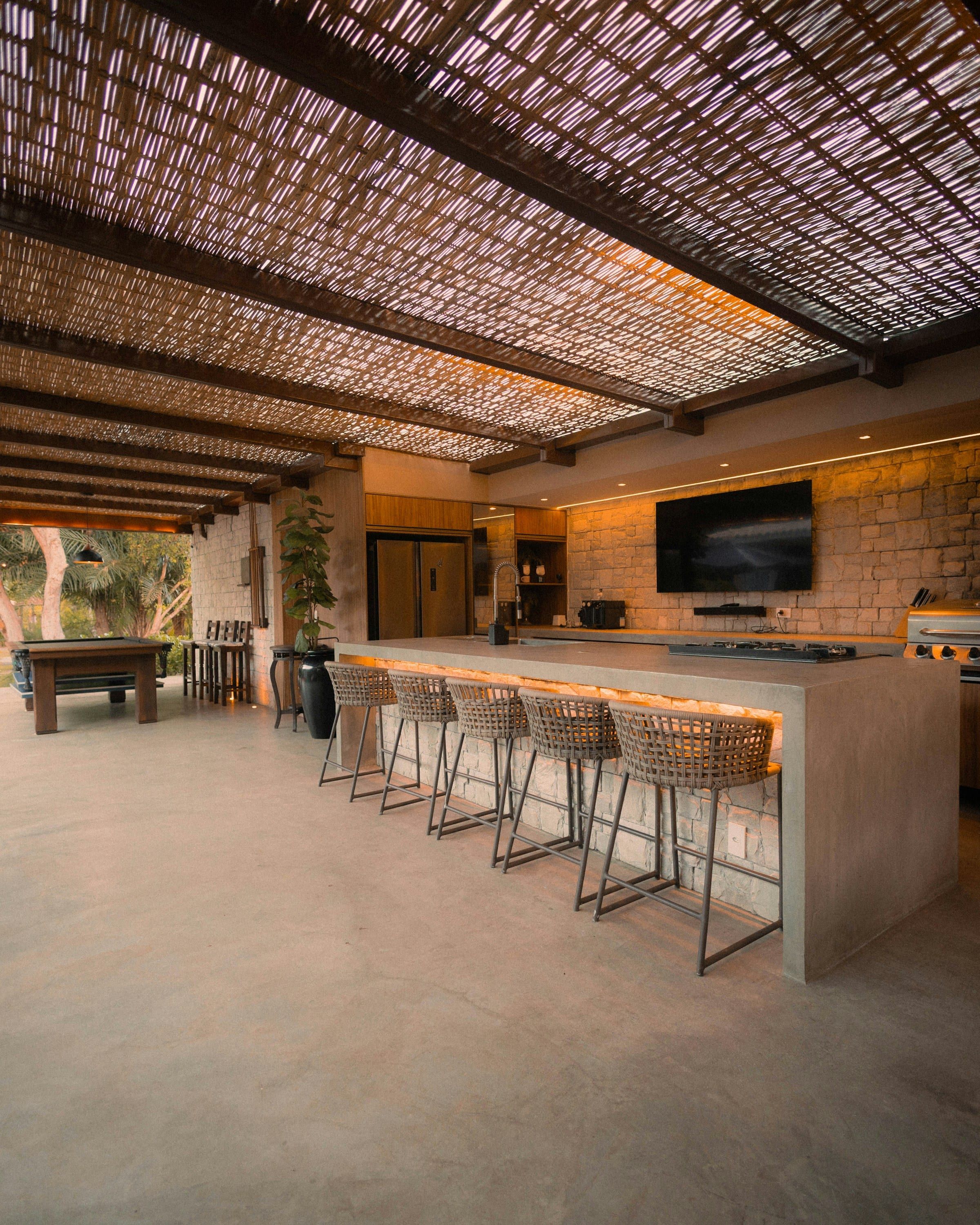 Covered outdoor kitchen with a long bar, bar stools and a pool table under a slatted roof