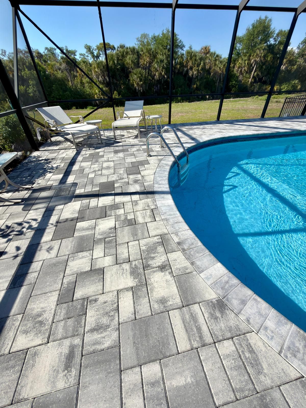 Gray paver pool deck with lounge chairs inside a screen enclosure, facing an open field