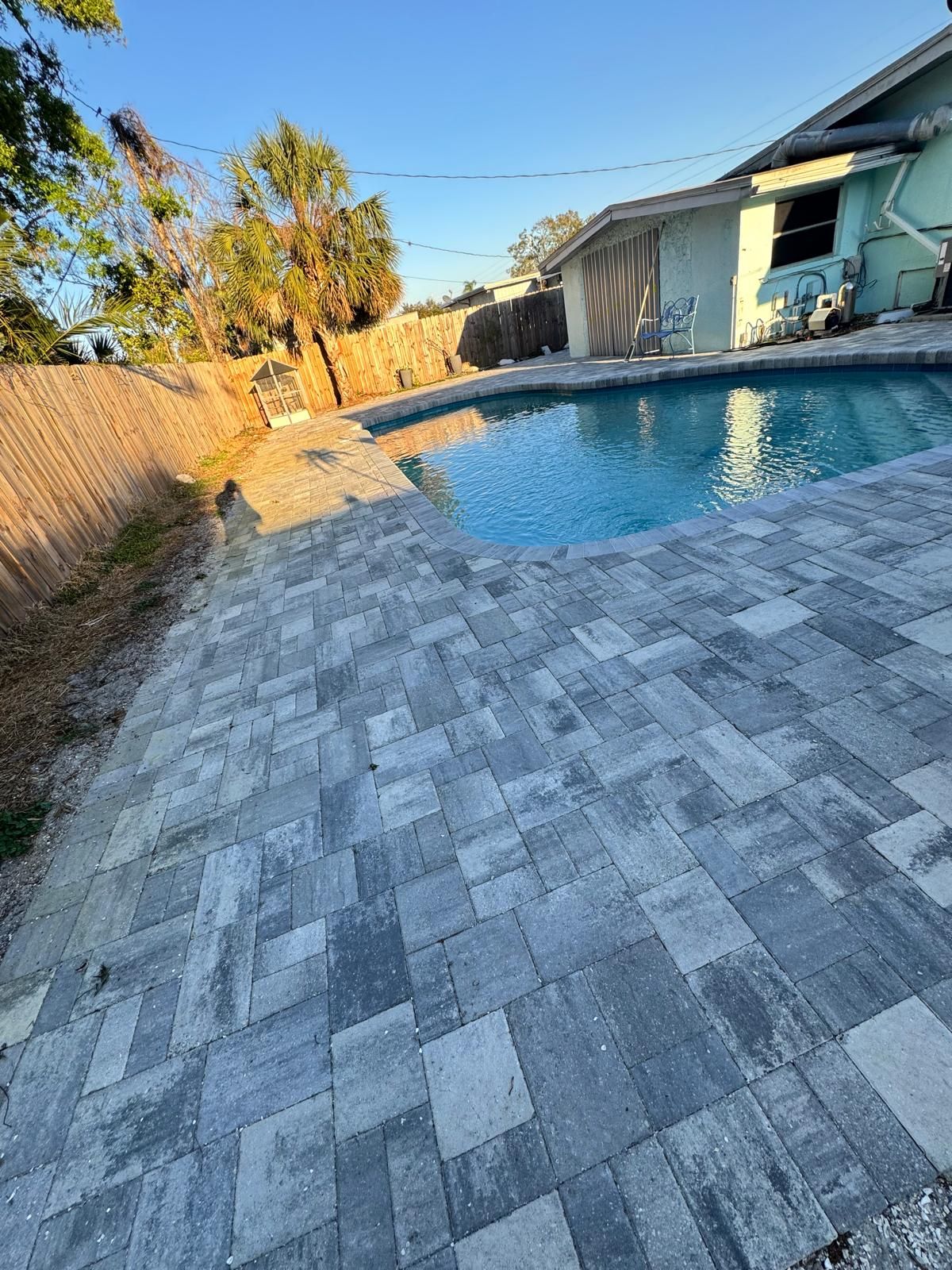 New gray paver deck around a kidney-shaped pool in a fenced backyard at sunset