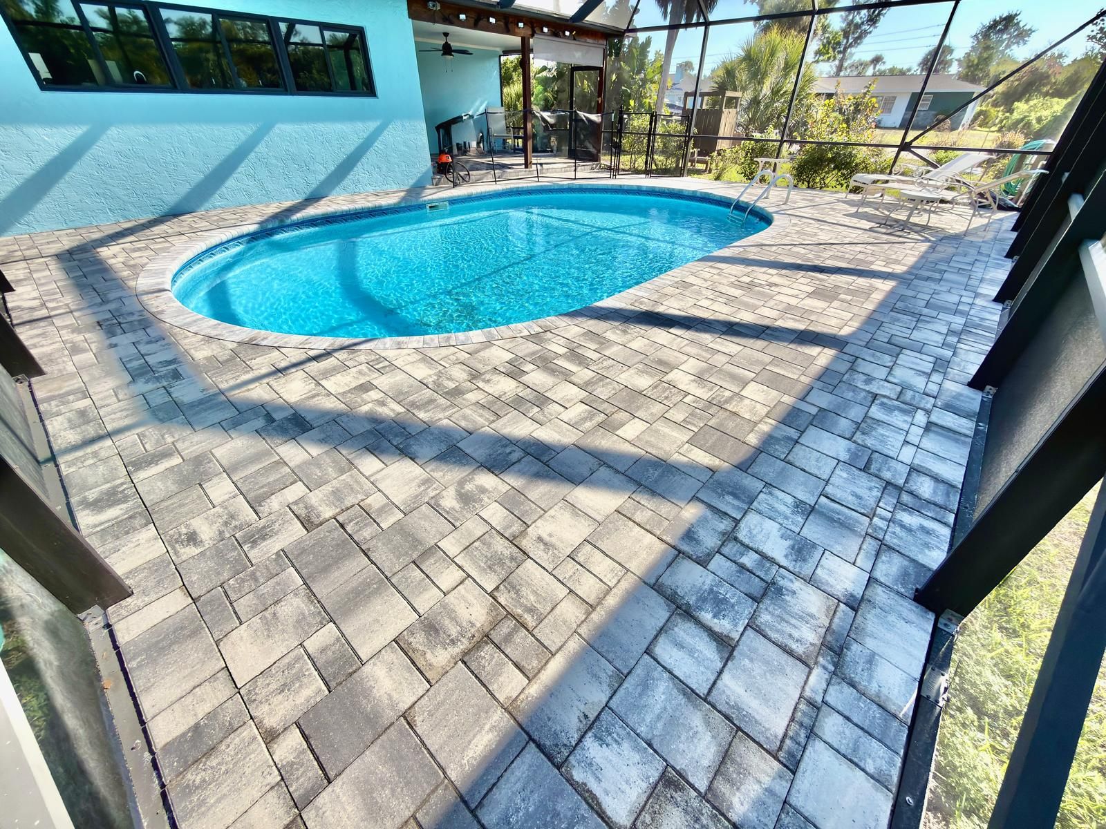 Gray and white paver deck around an oval pool inside a screen enclosure, next to a light blue house