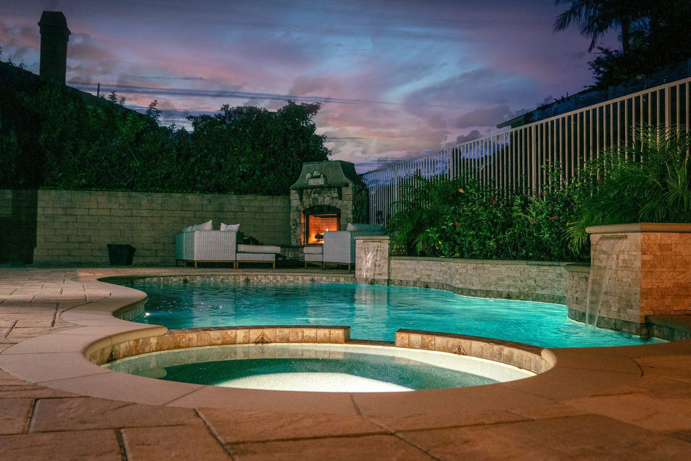 Lit spa spilling into a freeform pool at dusk, with a stone outdoor fireplace behind