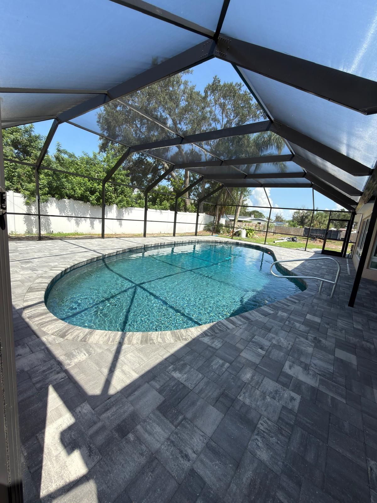 Gable-roof bronze screen enclosure over a freeform pool and a gray paver deck, with trees behind the fence