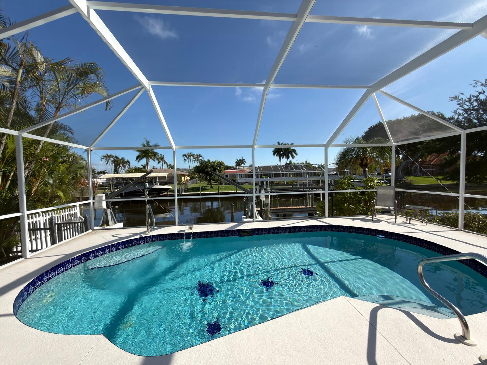 White aluminum screen enclosure over a kidney-shaped pool with blue tile accents, looking out to a canal and palm trees